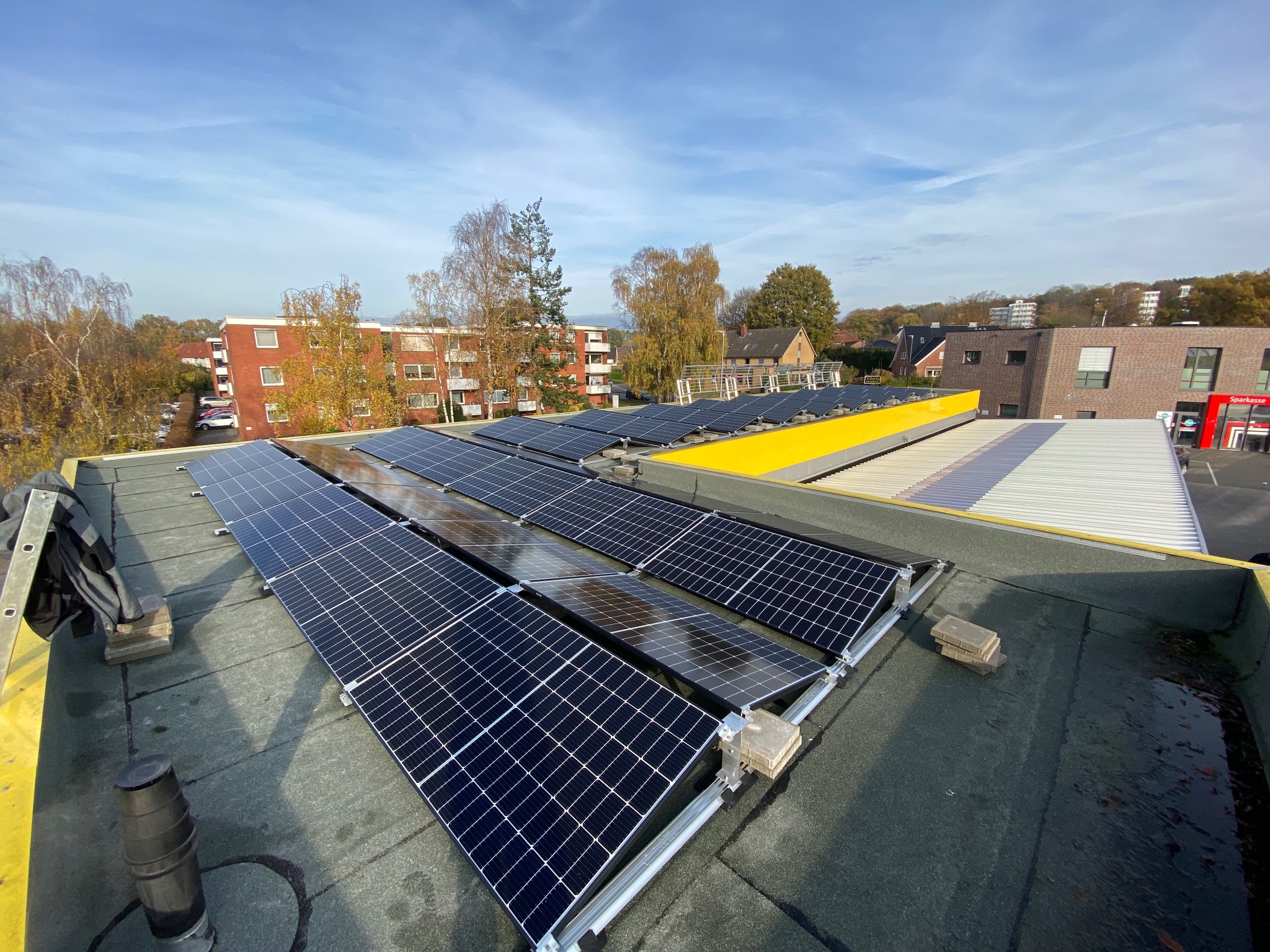 Instalacja paneli słonecznych na płaskim dachu budynku komercyjnego, widok z góry na panele w rzędach, na tle budynki mieszkalne i drzewa jesienią.