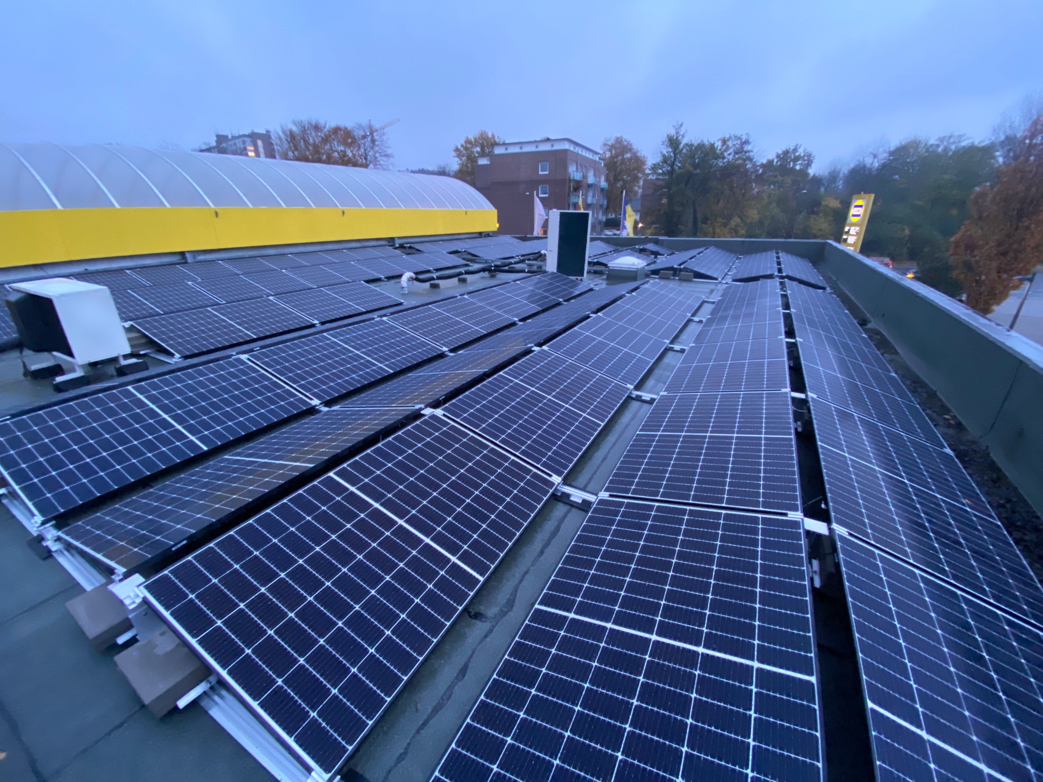 Rozległa instalacja paneli słonecznych na płaskim dachu budynku komercyjnego, widok z góry w pochmurny dzień, z widocznymi elementami montażowymi i sąsiednimi budynkami w tle.