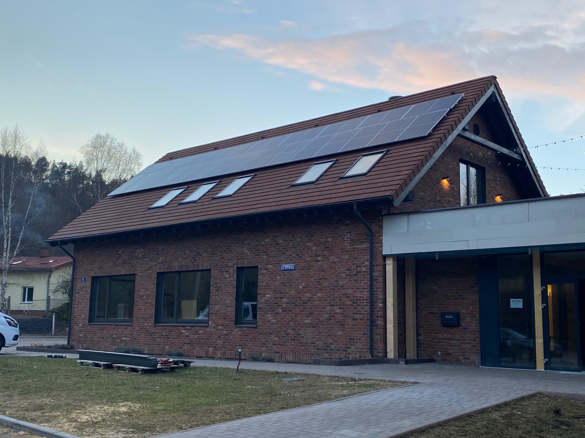 Dom z czerwonej cegły z panelami słonecznymi na dachu w Rozpędzinach, widoczne okna dachowe i fragment ulicy z zaparkowanym samochodem.