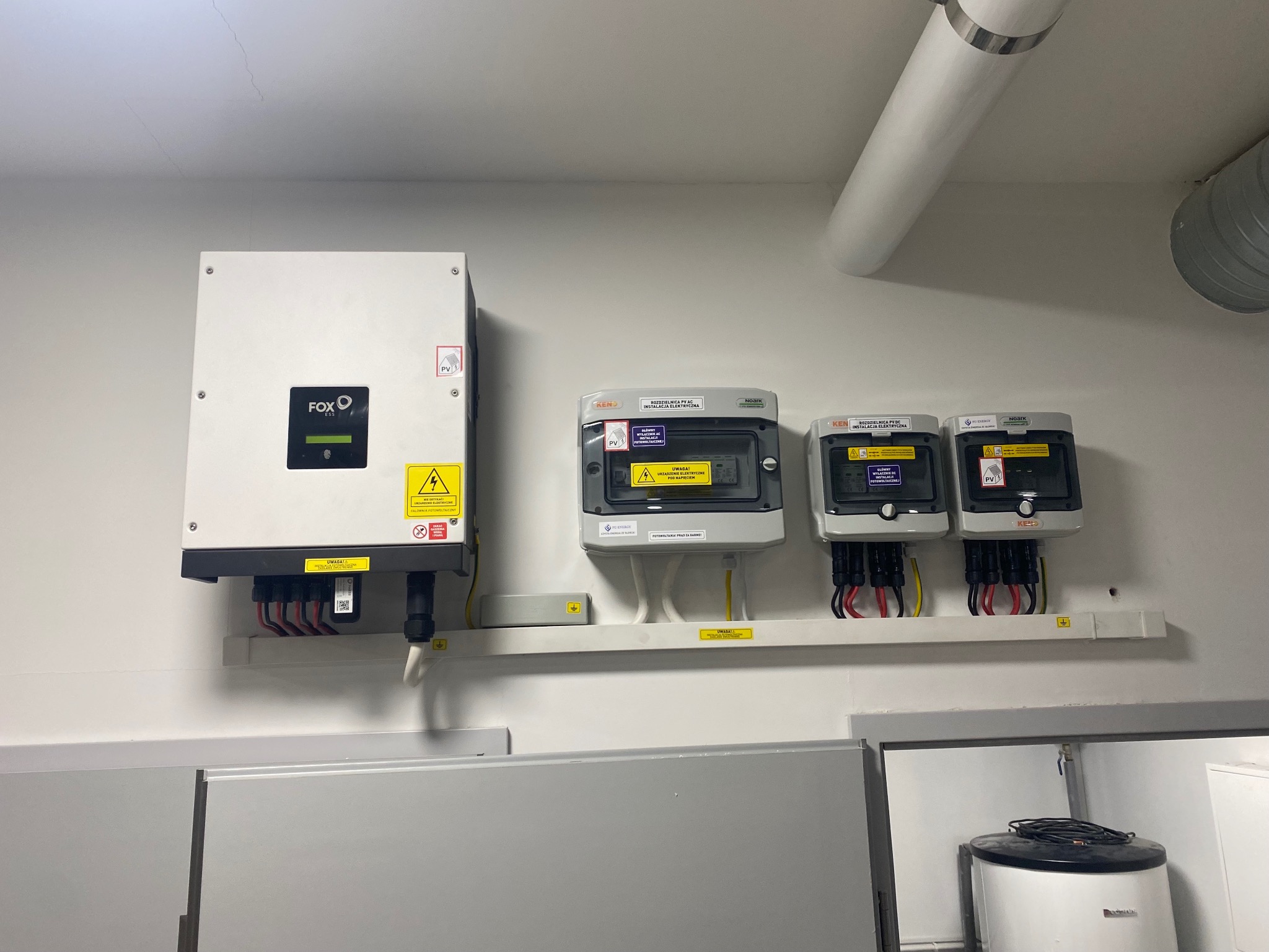 Instalacja elektryczna z falownikiem Fox i rozdzielnicami PV AC firm KEN i Noark, zamontowana na ścianie w pomieszczeniu technicznym z widocznym bojlerem.