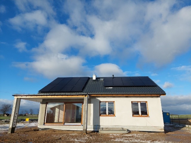 Dom jednorodzinny z zamontowanymi panelami fotowoltaicznymi na dachu, widok z boku, błękitne niebo z chmurami w tle.