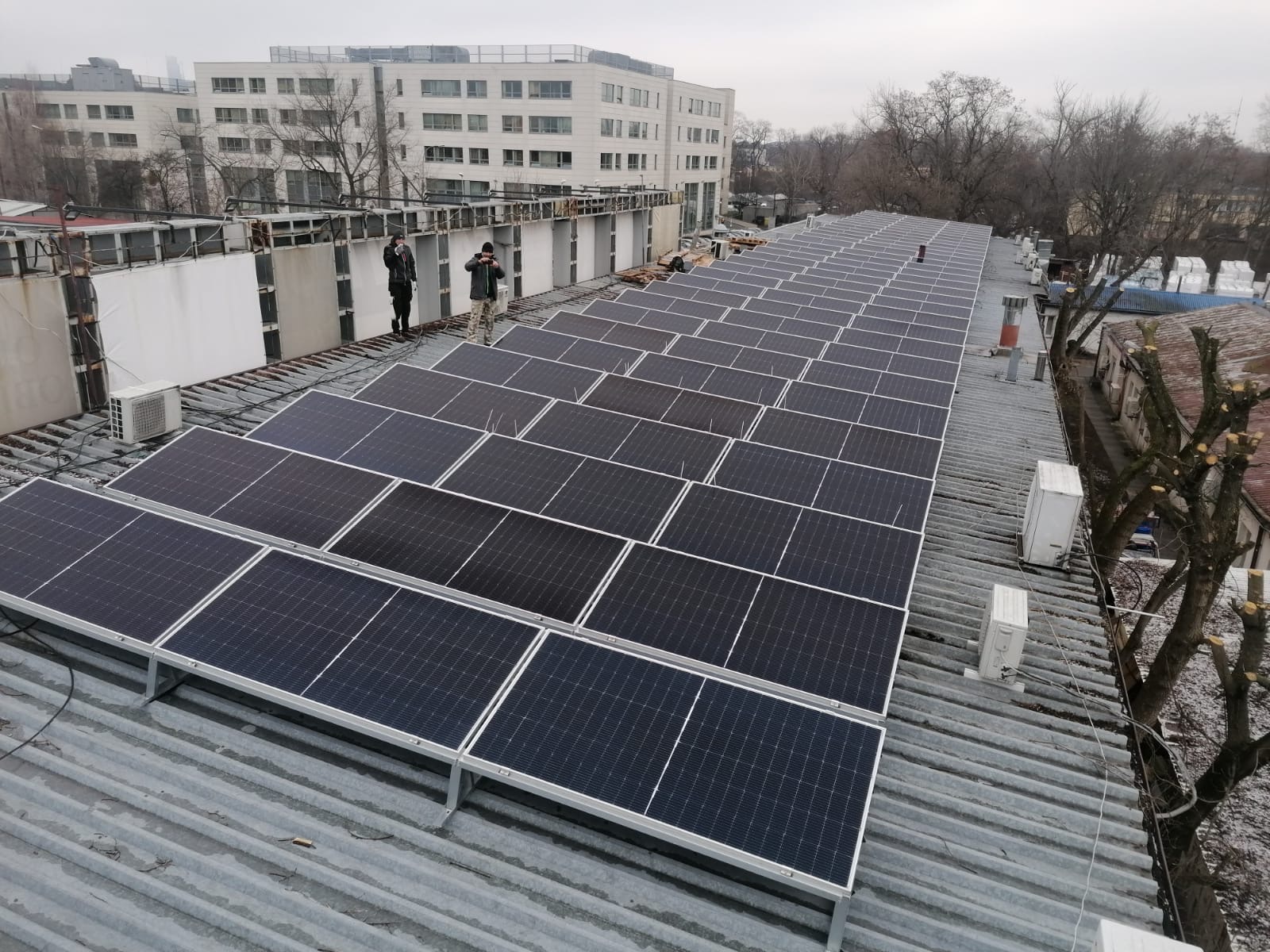 Instalacja paneli słonecznych na falistym dachu z blachy, widoczni dwaj instalatorzy w oddali, budynek mieszkalny w tle.