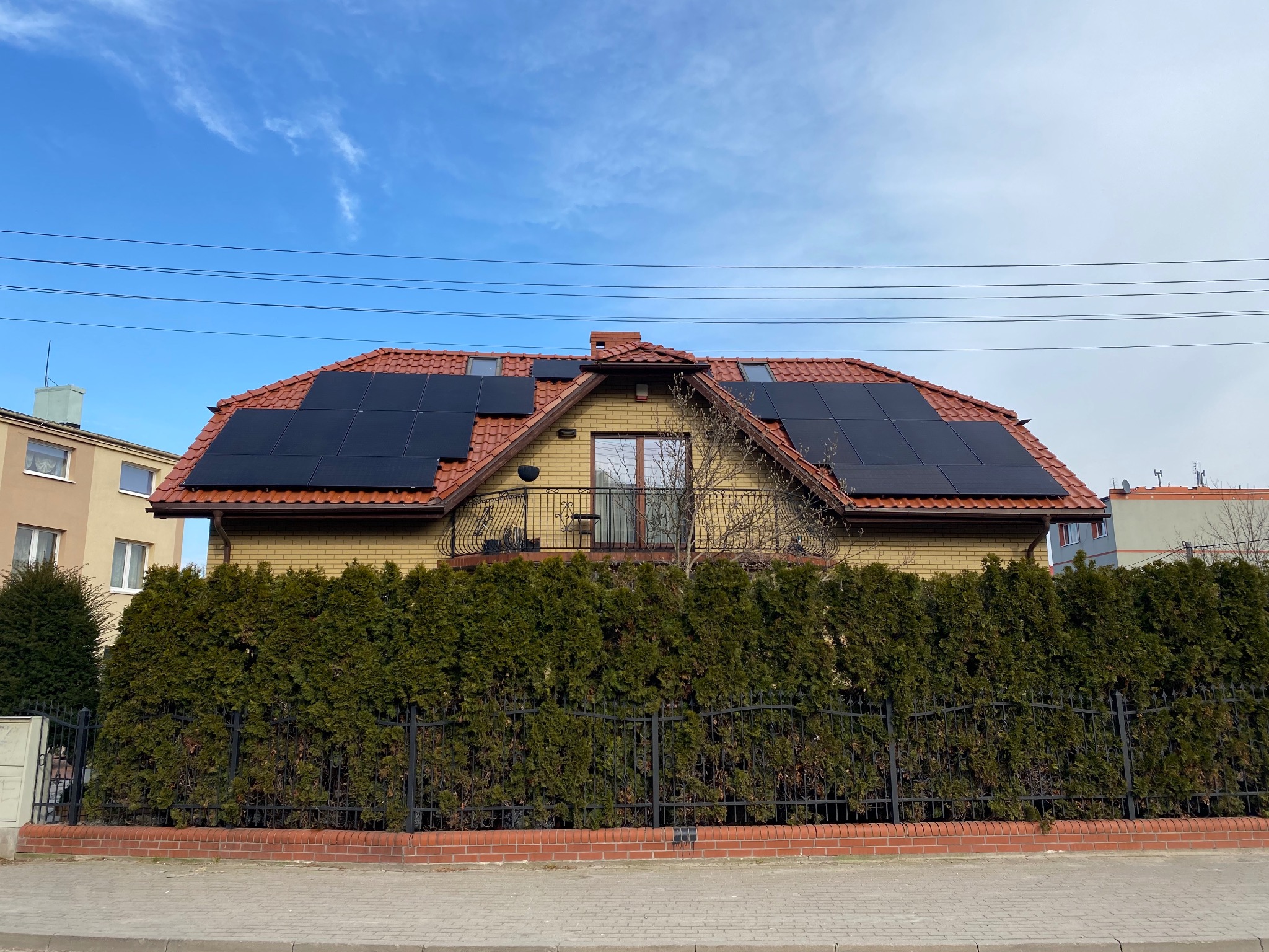 Dom jednorodzinny z panelami słonecznymi na dachu z czerwonej dachówki, widok z poziomu ulicy, zieleń żywopłotu w tle