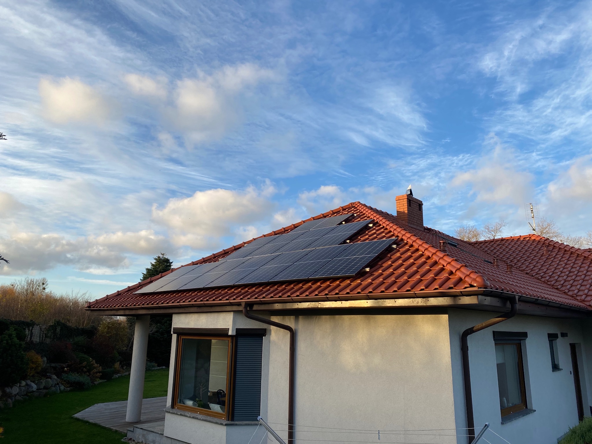 Panele słoneczne na dachu domu z czerwoną dachówką, błękitne niebo z chmurami w tle.