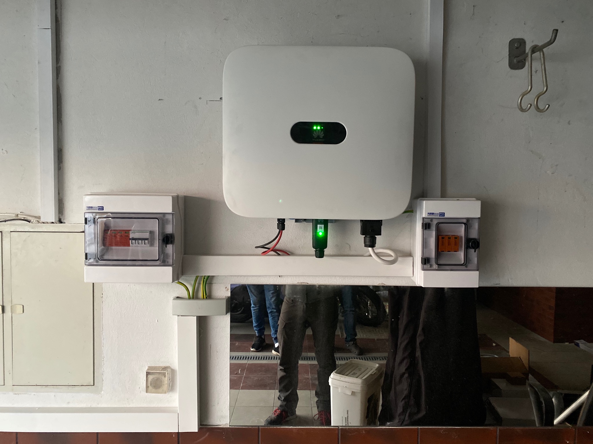 Biały inwerter z zielonymi diodami LED zamontowany na ścianie, obok skrzynki z bezpiecznikami. W lustrzanym odbiciu widoczne osoby i elementy otoczenia.