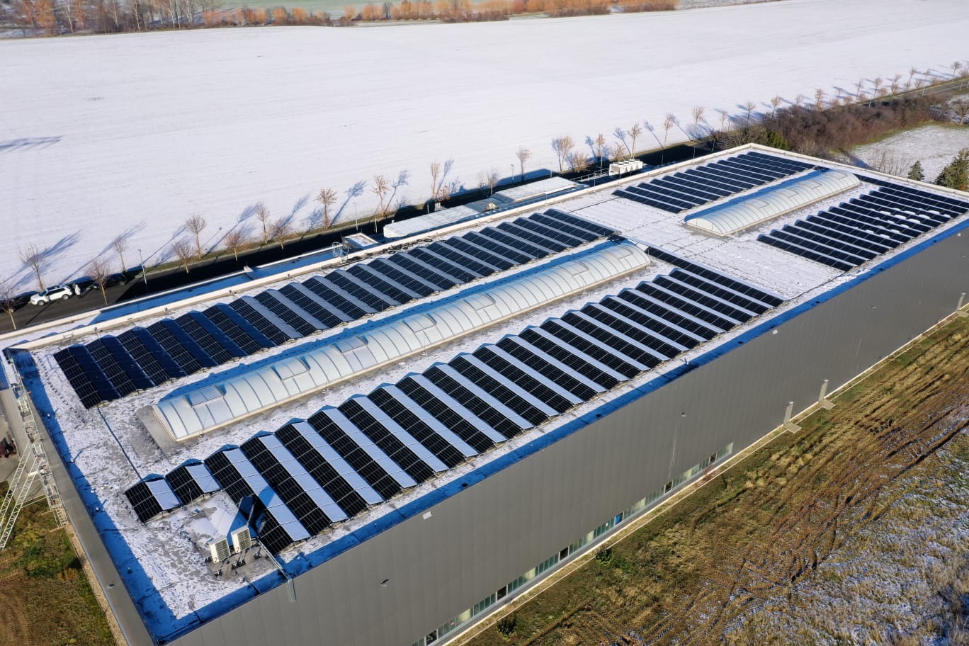 Widok z góry na duży budynek przemysłowy z panelami słonecznymi na dachu, częściowo pokrytymi śniegiem, w tle ośnieżone pole i drzewa.