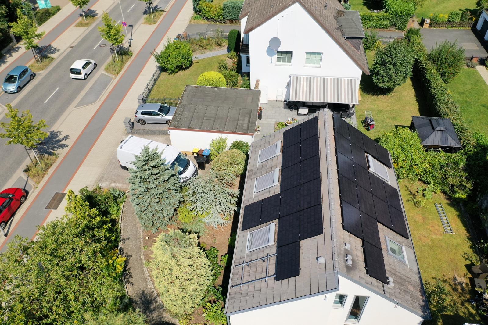 Widok z góry na dom z panelami słonecznymi na dachu, biały van zaparkowany obok, zadbany ogród i ulica z samochodami w tle.