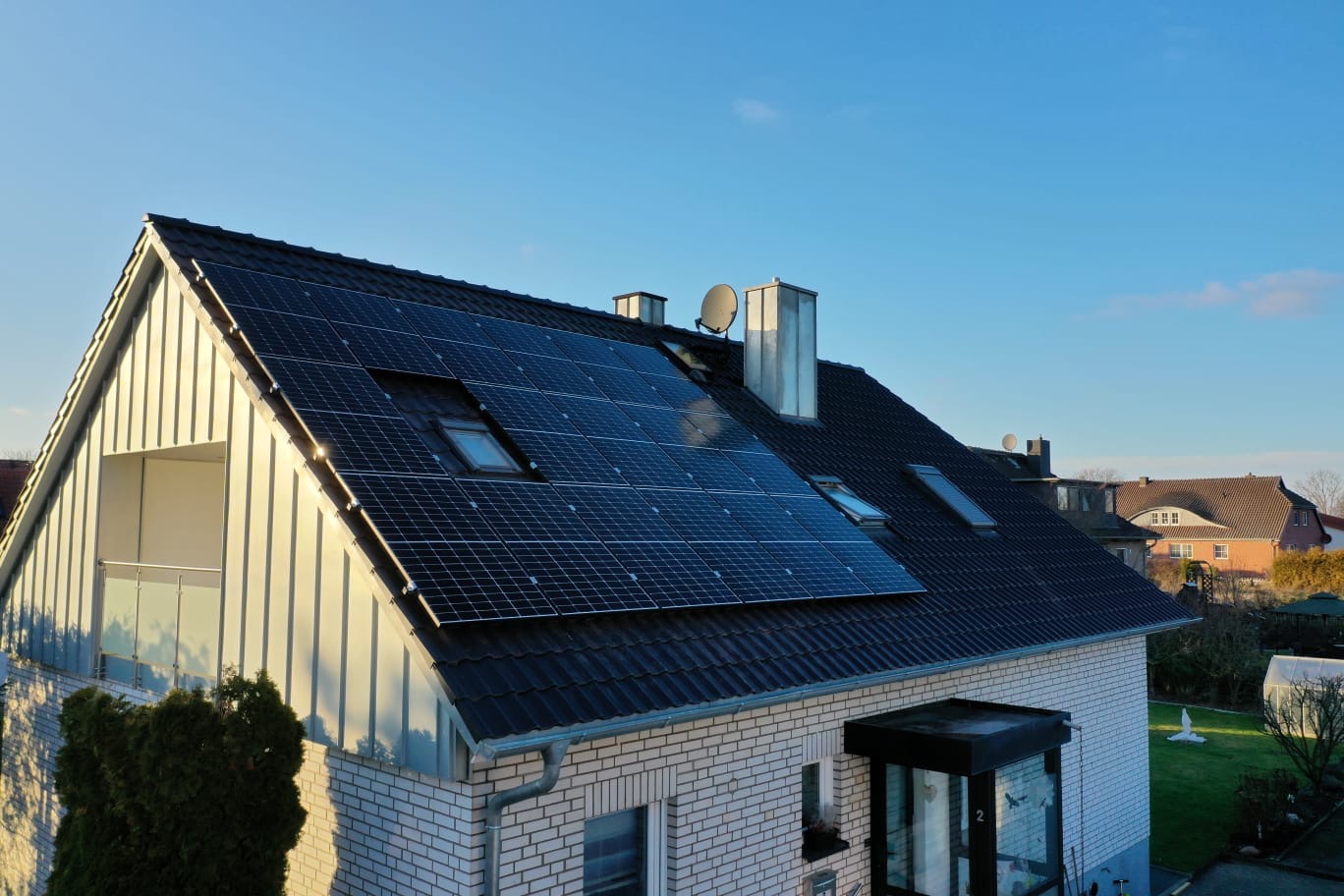 Instalacja fotowoltaiczna na dachu domu jednorodzinnego z białej cegły, widoczne okna dachowe, komin i antena satelitarna, w tle błękitne niebo.