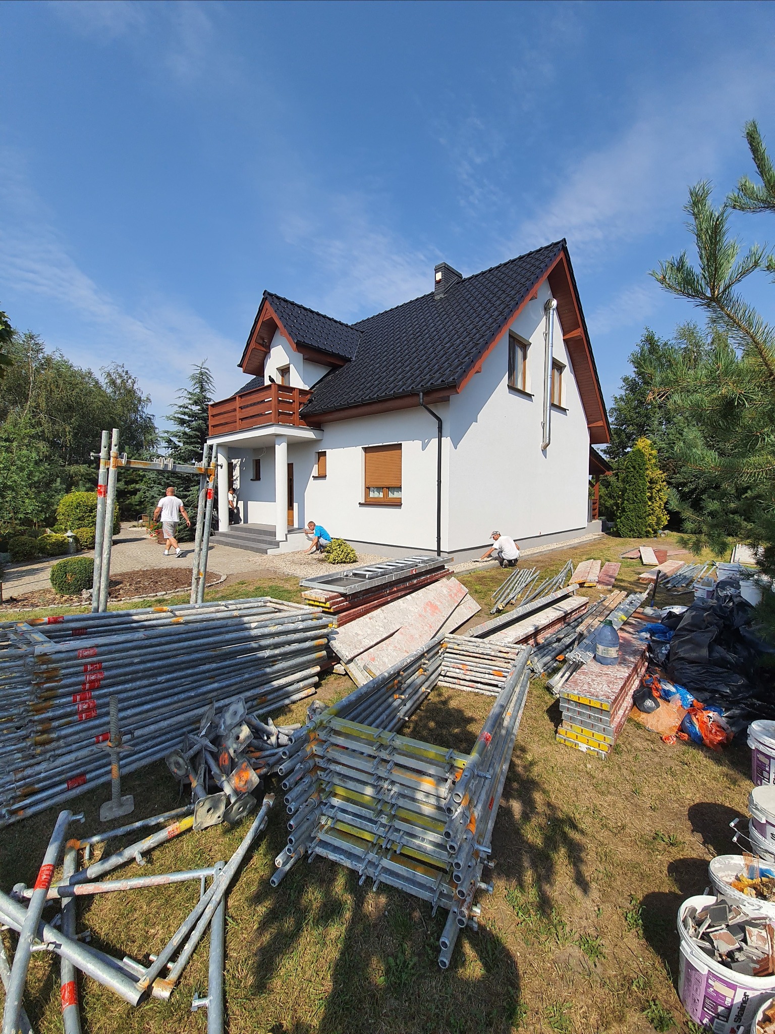 Dom jednorodzinny w trakcie prac elewacyjnych: widoczne rusztowania, materiały budowlane i pracownicy w trakcie wykonywania drobnych poprawek przy elewacji.