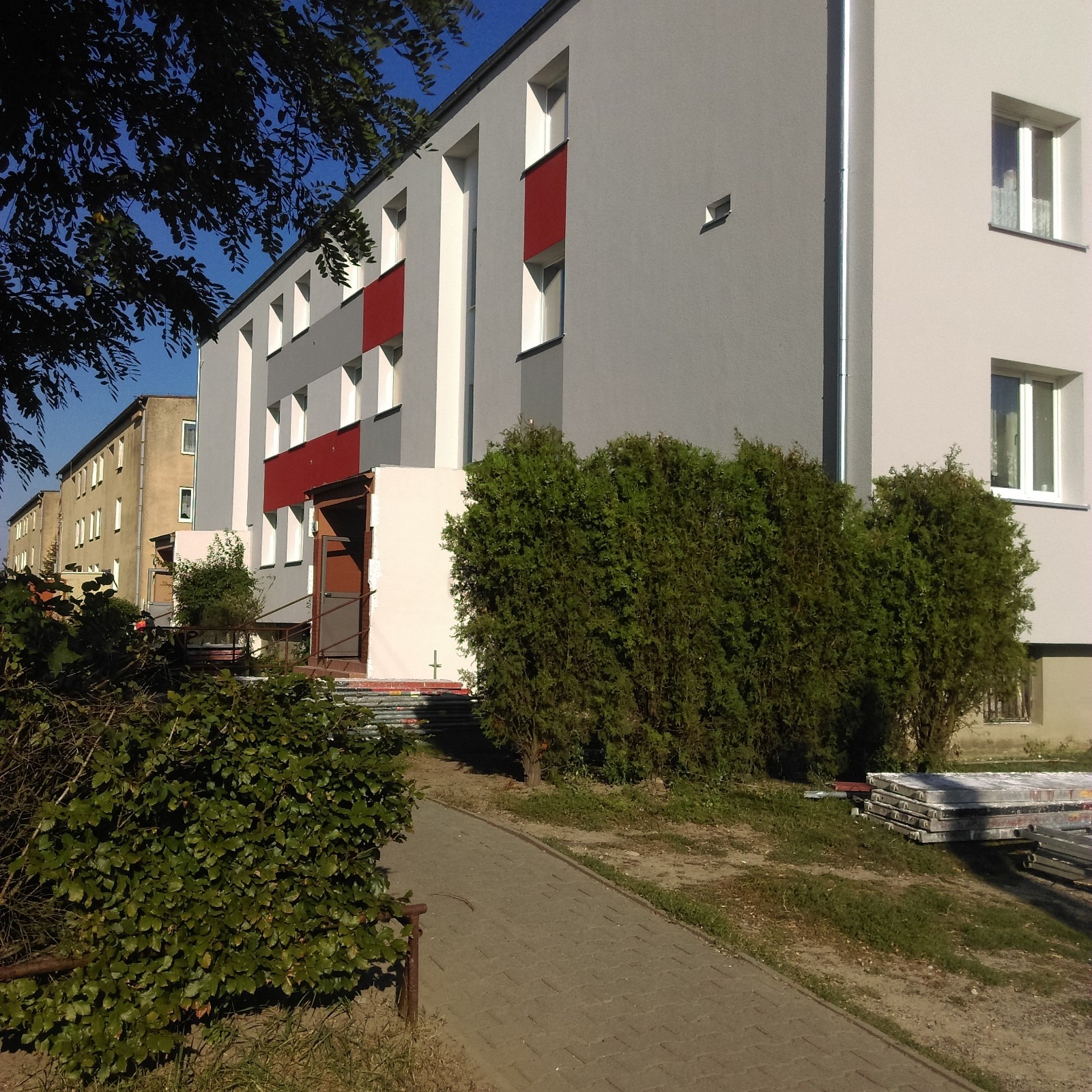 Odnowiona elewacja budynku wielorodzinnego w odcieniach szarości, bieli i czerwieni, widoczna z poziomu chodnika, z fragmentem zieleni w kadrze i rusztowaniem w tle.