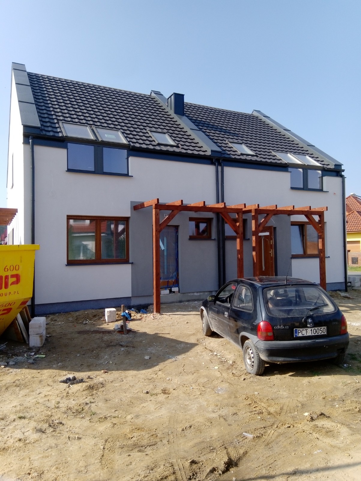 Nowoczesny dom bliźniak po remoncie elewacji, z szarą fasadą, ciemną dachówką, drewnianą pergolą i zaparkowanym czarnym samochodem osobowym.