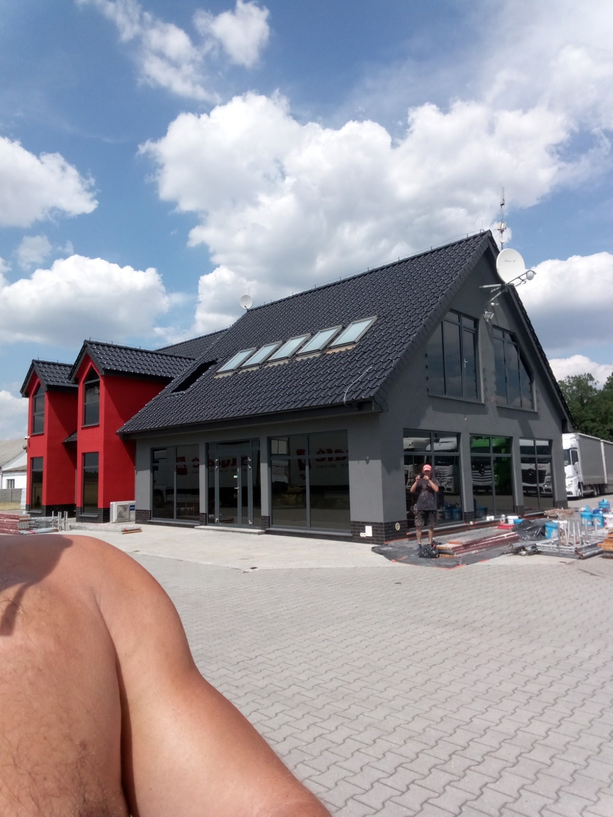 Nowoczesny budynek po renowacji elewacji, łączący szarą i czerwoną kolorystykę, z licznymi oknami i satelitami na dachu, częściowo widoczny fragment sylwetki fotografa w dolnym lewym rogu.