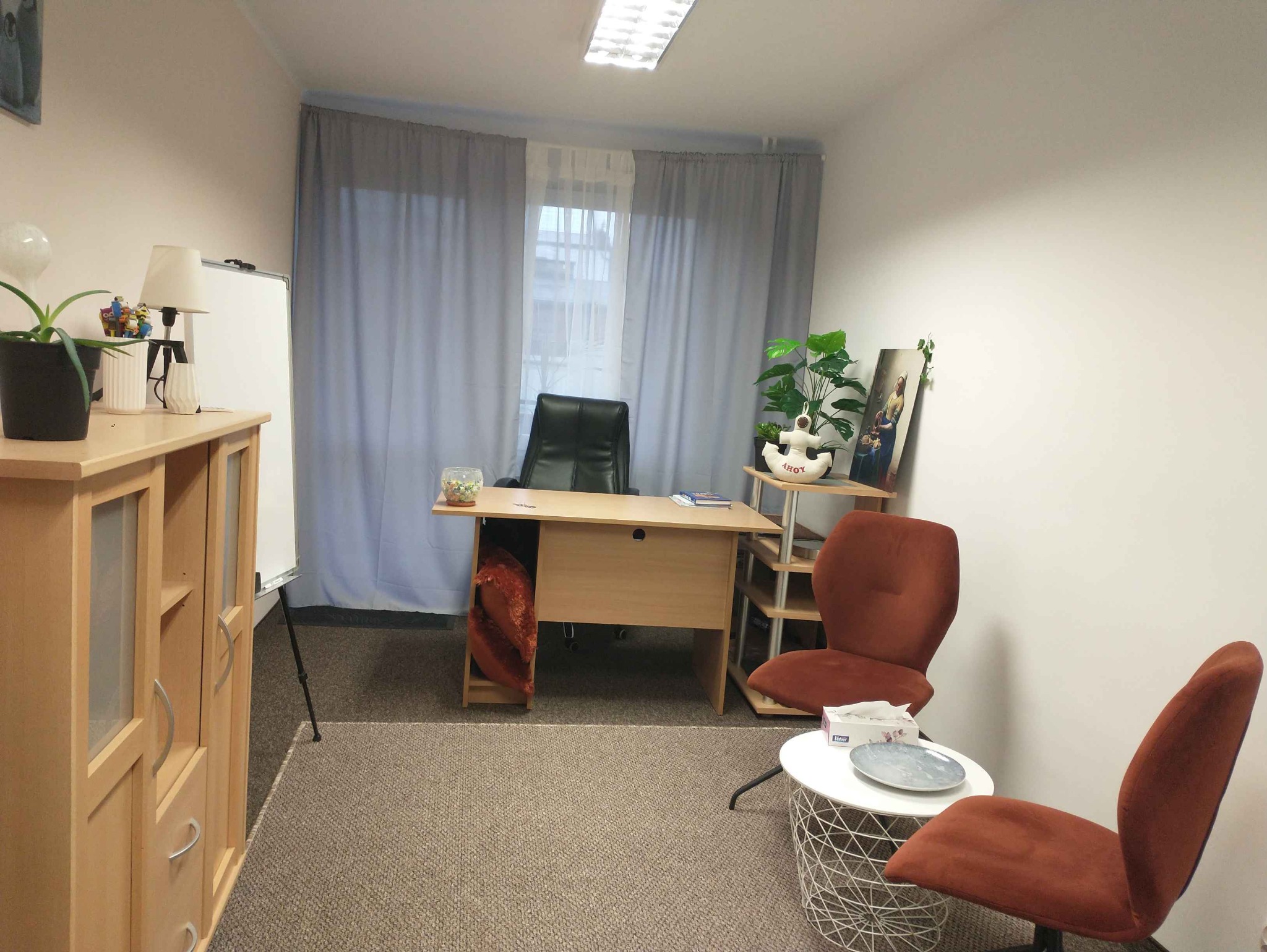 Gabinet z biurkiem, krzesłami w kolorze ceglastym, kwiatami doniczkowymi i tablicą suchościeralną, utrzymany w neutralnej kolorystyce.