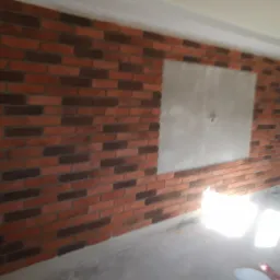 Ściana wykończona cegłą klinkierową w odcieniach brązu i czerwieni, z widocznym otworem rewizyjnym zakrytym płytą gipsową, w tle widoczny kubeł z farbą.