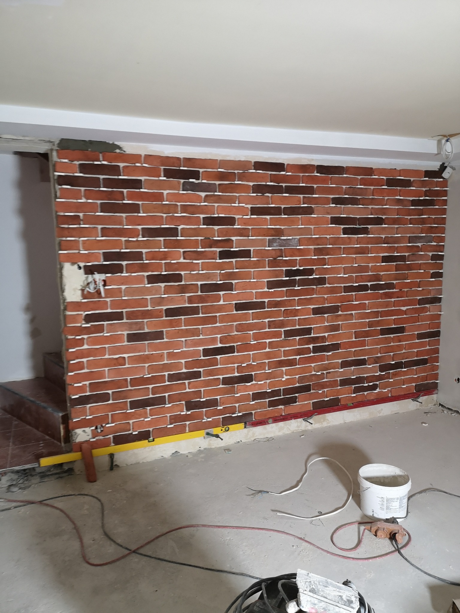 Ściana wykończona cegiełkami dekoracyjnymi z fugą, widoczne narzędzia i materiały budowlane na podłodze w trakcie prac wykończeniowych.