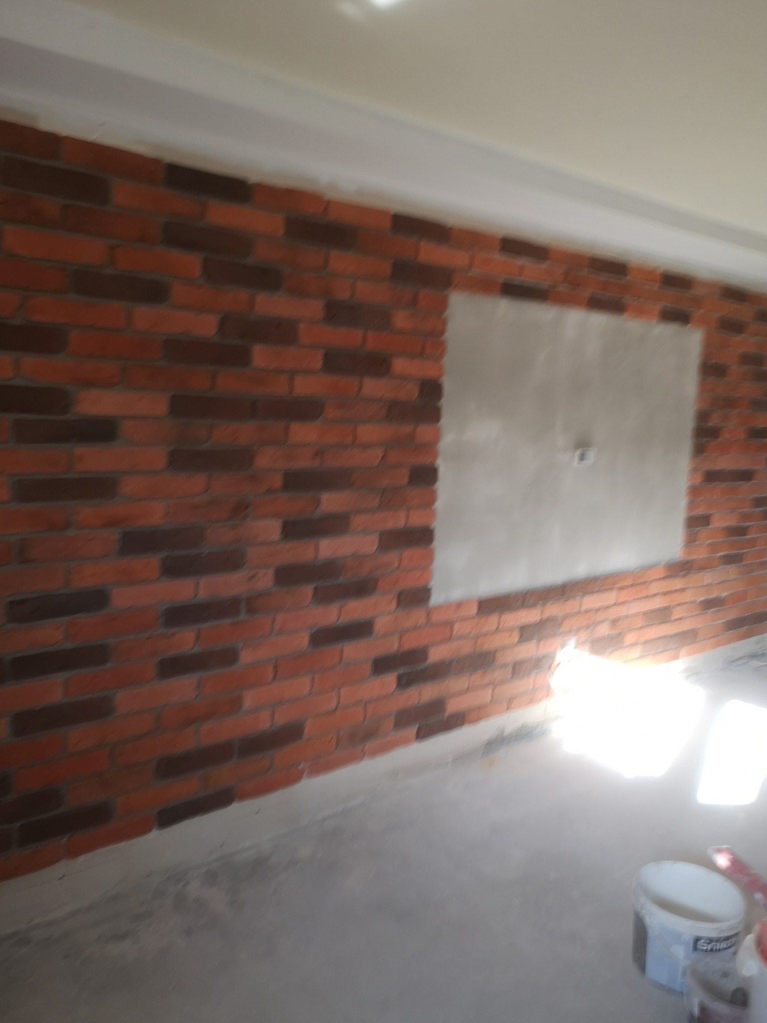 Ściana wykończona cegłą klinkierową w odcieniach brązu i czerwieni, z widocznym otworem rewizyjnym zakrytym płytą gipsową, w tle widoczny kubeł z farbą.