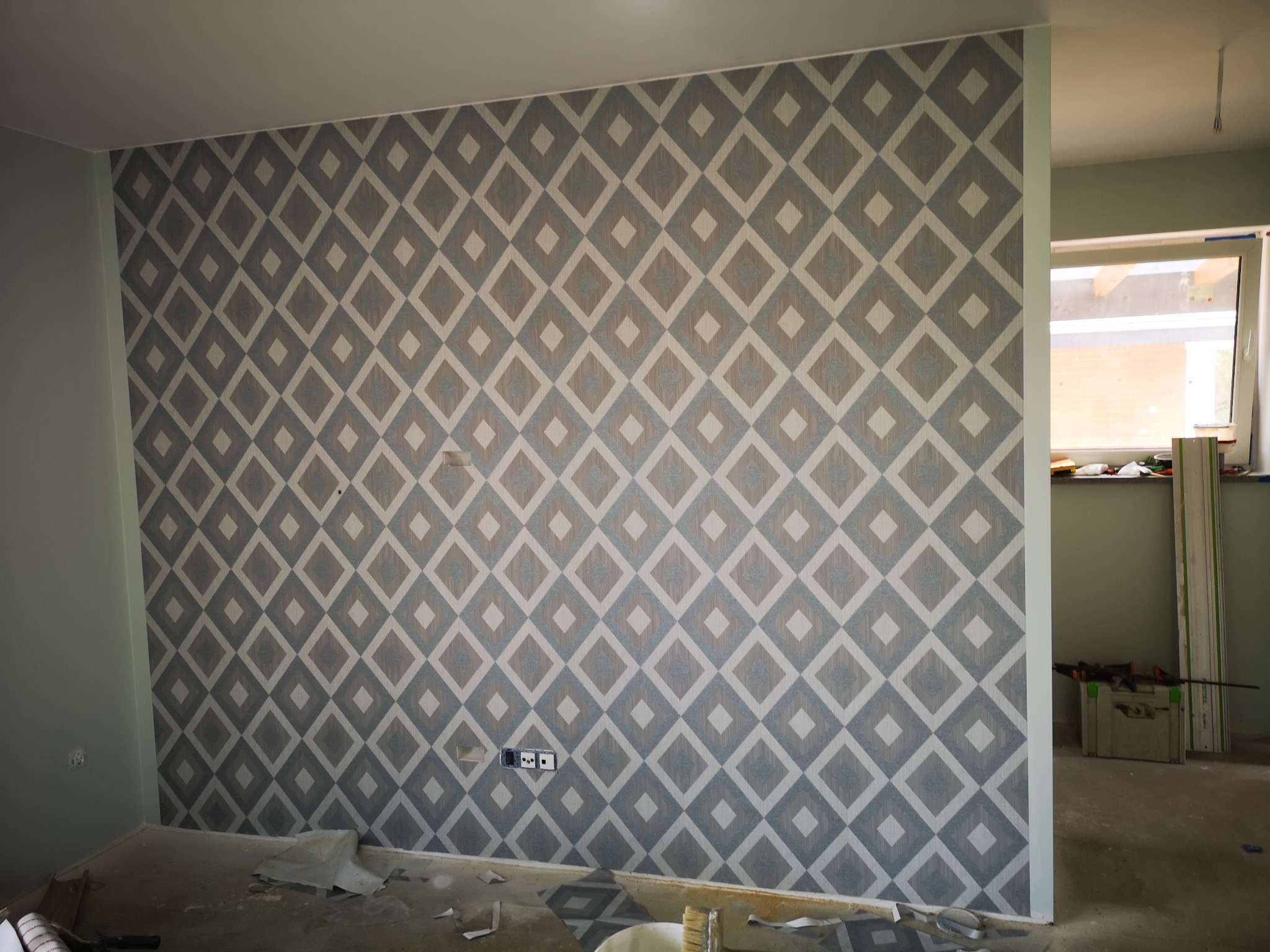 Ściana pokryta tapetą w geometryczny wzór rombów w odcieniach szarości i bieli, widoczne gniazdka elektryczne i fragment podłogi z resztkami tapety, w tle widoczne okno i narzędzia.