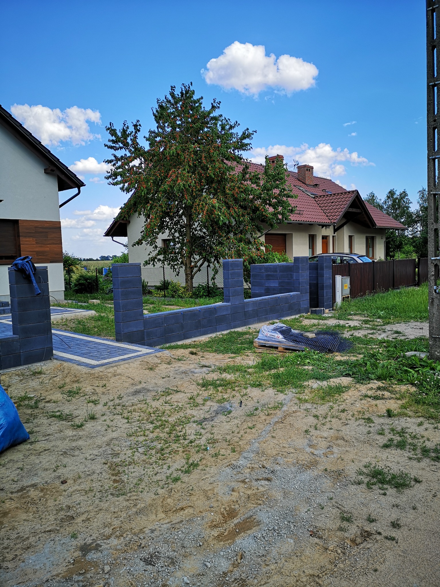Wznoszenie ogrodzenia z prefabrykowanych, grafitowych bloczków betonowych na posesji z widocznymi dwoma budynkami mieszkalnymi i drzewem owocowym.