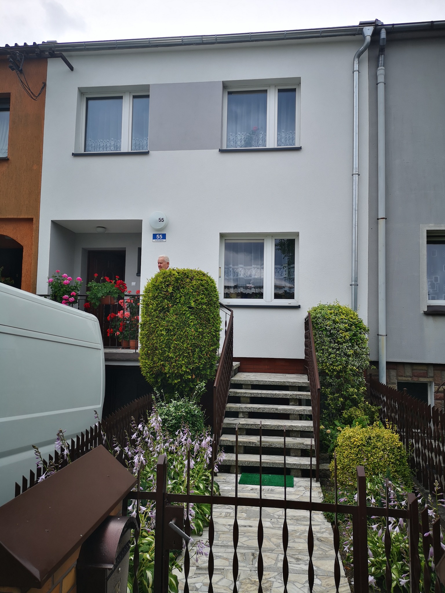 Dwupiętrowy dom szeregowy z betonowymi schodami prowadzącymi do wejścia, ozdobiony roślinami w donicach na balkonie i przy schodach, widoczny fragment białego samochodu dostawczego.