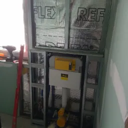 Instalacja podtynkowa WC w stelażu z profili stalowych, widoczna izolacja termiczna na ścianie przy oknie, poziomica i wkrętarka obok, prace w toku w Wydminach.