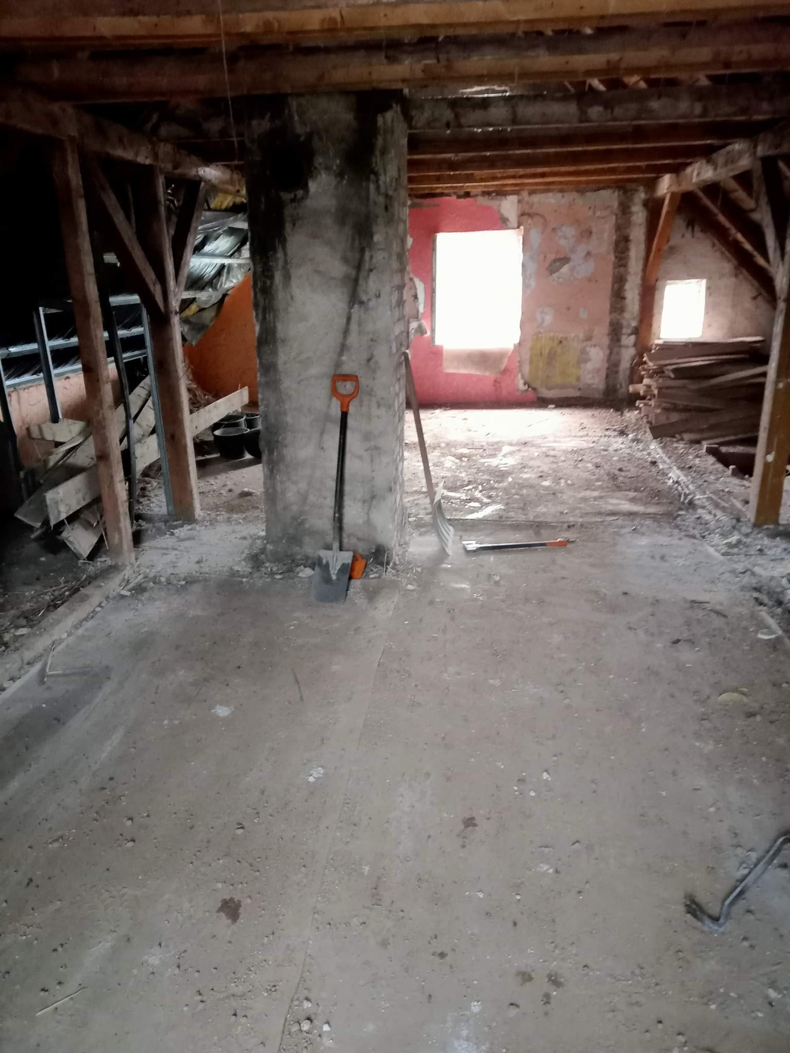 Poddasze w trakcie demontażu, widoczne drewniane belki, ceglany komin z opartą o niego łopatą, resztki tynku na ścianach i narzędzia na podłodze.