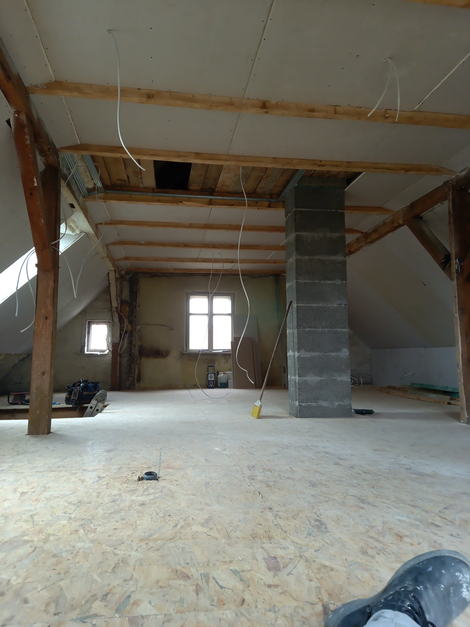 Poddasze w trakcie prac wykończeniowych: widoczne drewniane belki stropowe, częściowo zamontowana płyta gipsowa na suficie, murowany komin z bloczków, wystające przewody elektryczne i fragment...