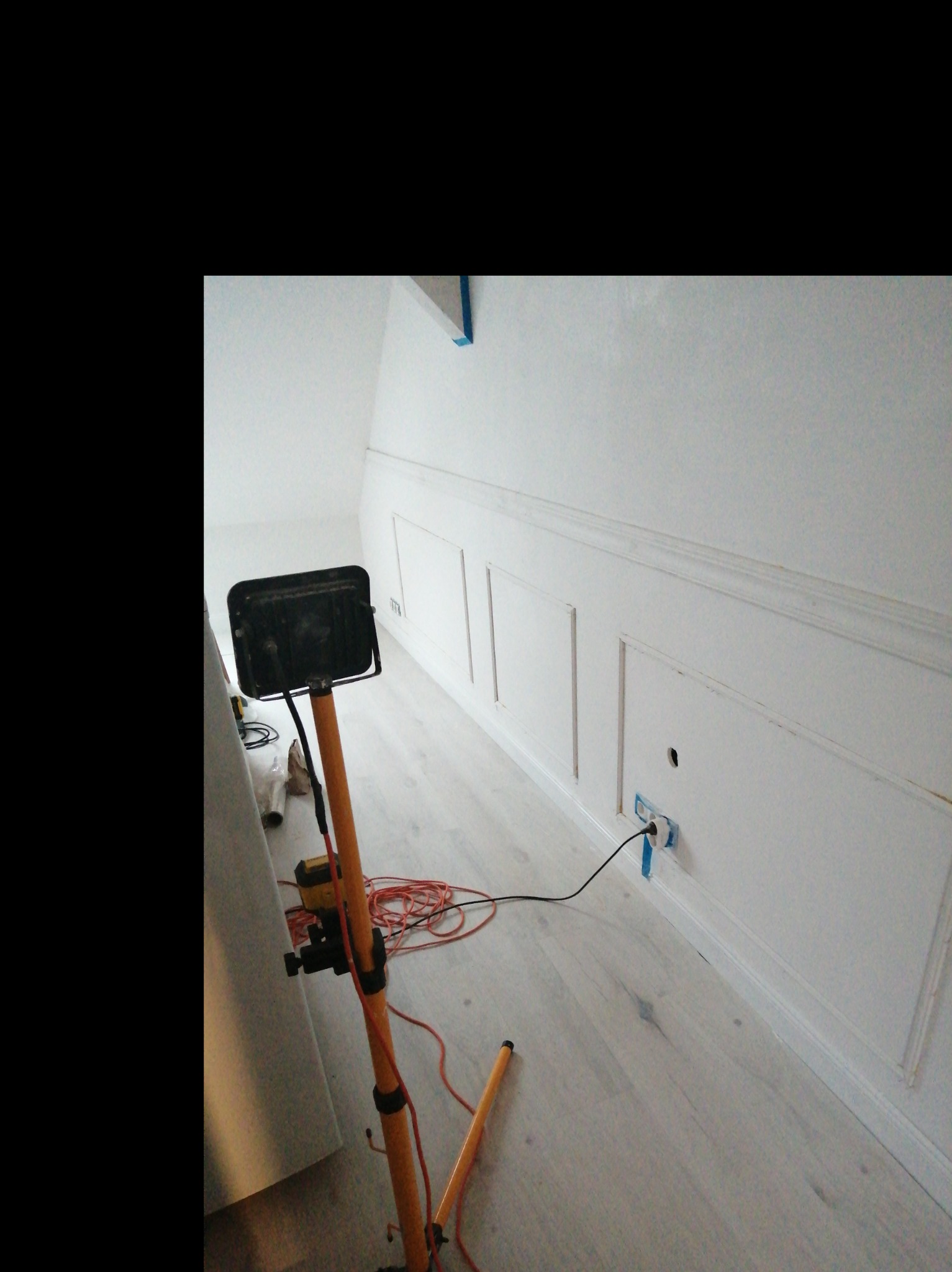 Biała ściana z panelami dekoracyjnymi i listwami przypodłogowymi, oświetlona lampą roboczą na trójnogu, widoczny kabel zasilający i gniazdko zabezpieczone taśmą malarską.