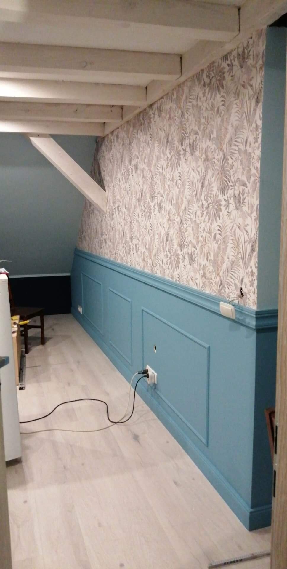 Pokój na poddaszu w trakcie wykańczania: ściana z tapetą w roślinne wzory, niebieska boazeria z panelami, białe belki stropowe, widoczne gniazdka elektryczne i kabel na podłodze.