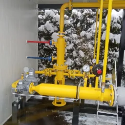 Żółta instalacja gazowa z manometrami i zaworami, na tle ośnieżonych krzewów, wewnątrz metalowej obudowy.