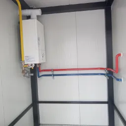 Biały piec gazowy zamontowany na ścianie z widocznymi rurami gazowymi (żółta) oraz rurami doprowadzającymi wodę (czerwona i niebieska) w pomieszczeniu o metalowej konstrukcji.