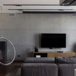 Nowoczesny salon z szarą ścianą z paneli imitujących beton, wiszącym fotelem-bańką na łańcuchu, szarą sofą i drewnianą podłogą.