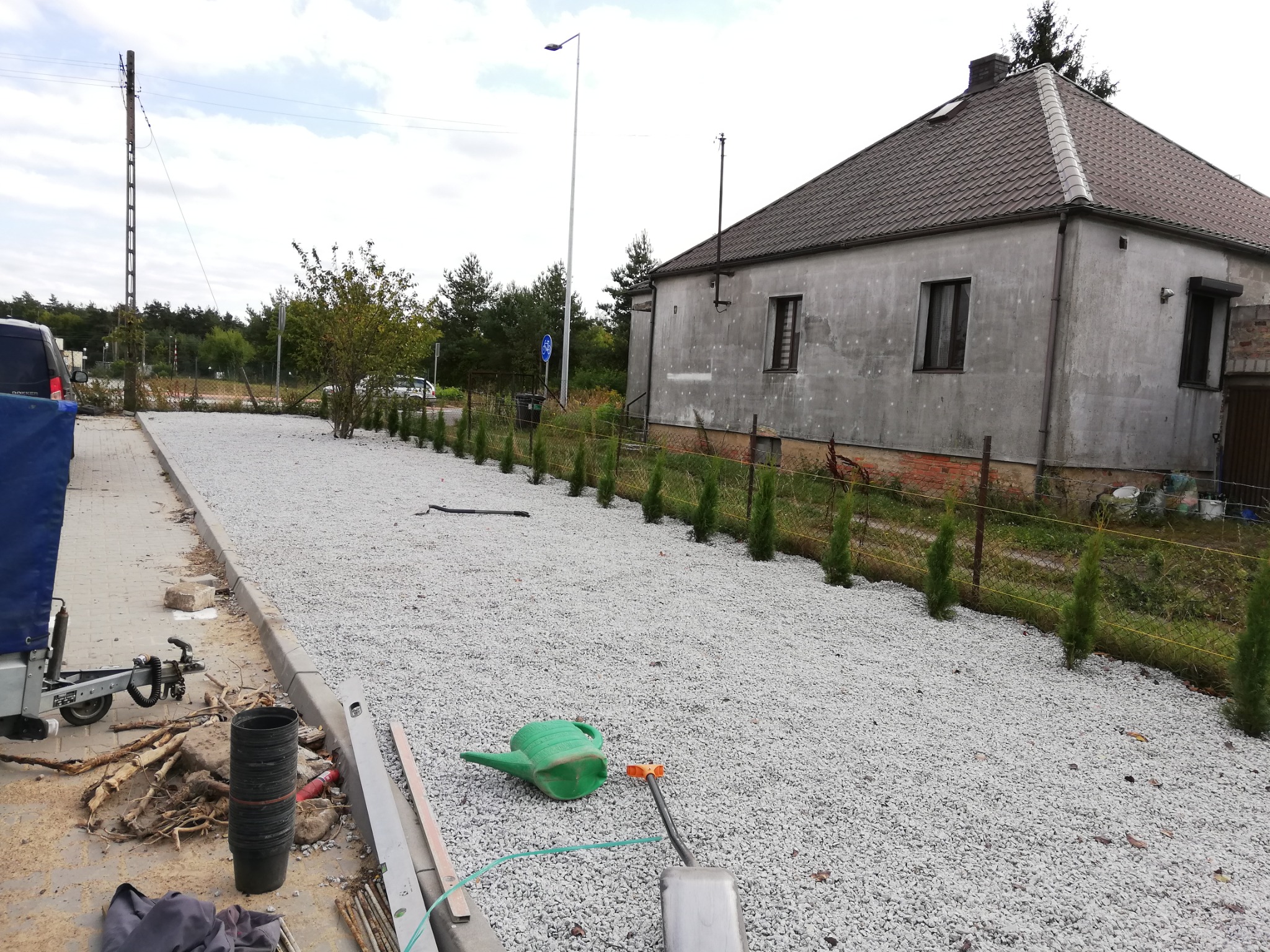 Wykończony podjazd z białego kruszywa obok szarego domu, rząd młodych tuj wzdłuż ogrodzenia, zielona konewka i narzędzia na podłożu.