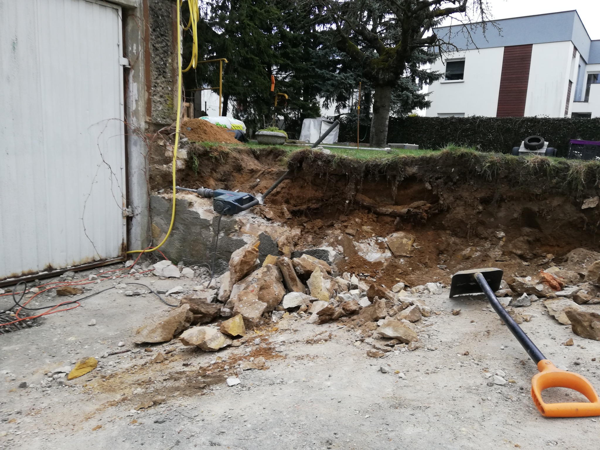 Wykop w ziemi z widocznym gruzem, narzędziami (szpadel, młot udarowy) i kablem elektrycznym, garaż w tle.