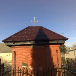 Ceglana kapliczka z brązowym dachem z blachy trapezowej, zwieńczona metalowym krzyżem, widoczna na tle błękitnego nieba.