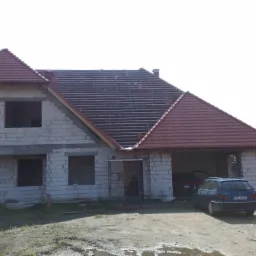 Niedokończony dom w trakcie budowy z częściowo pokrytym dachem czerwoną dachówką, widoczne krokwie bez pokrycia na pozostałej części dachu, szare ściany z pustaków, samochód osobowy zaparkowany...