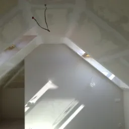 Poddasze ze skośnym sufitem, częściowo pokryte płytami kartonowo-gipsowymi, widoczne przewody elektryczne i światło słoneczne wpadające przez okno dachowe.