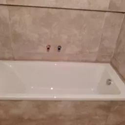 Prostokątna, biała wanna w obudowie z beżowych płytek, z widocznymi zaworami ciepłej i zimnej wody.