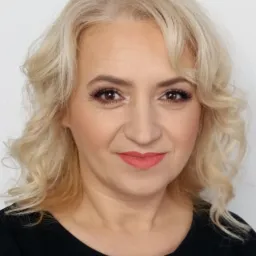 Portret kobiety z blond kręconymi włosami, delikatnym makijażem i czarną bluzką z suwakiem.