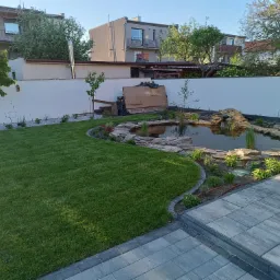 Oczko wodne z kamienną obudową i bujną roślinnością, otoczone trawnikiem z rolki i brukowanym chodnikiem. Biały mur w tle, drzewa i budynki mieszkalne.