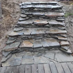 Schody z nieregularnych, płaskich kamieni w ogrodzie, prowadzące w górę zbocza, z drewnianym podestem u dołu, w tle ziemia i roślinność.