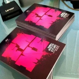 Stosy świeżo wydrukowanych, kwadratowych, czarno-r&oacute;żowych materiał&oacute;w reklamowych z datą 9.17.2011 leżą na biurku obok elektroniki.