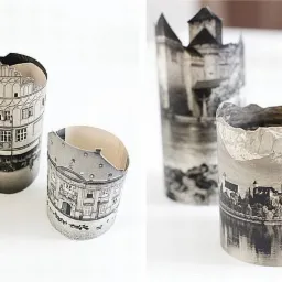 Trzy cylindryczne, papierowe modele budynków i krajobrazów w odcieniach szarości, z nierównymi krawędziami, ustawione na białym tle. Jeden model przedstawia kamienicę, drugi zamek nad jeziorem,...
