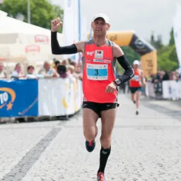 Biegacz z numerem 3 na koszulce, z uniesioną ręką, przekracza linię mety na brukowanej ulicy, w tle widoczni kibice i banery reklamowe.