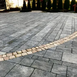 Ułożona kostka brukowa w odcieniach szarości z dekoracyjnym pasem jasnych kostek tworzącym łuk na podjeździe przed domem z tujami w tle.