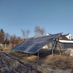 Panel słoneczny zamontowany na konstrukcji stalowej w ogrodzie, widoczny dom z białymi oknami i drabina oparta o konstrukcję, na ziemi ślady szronu.