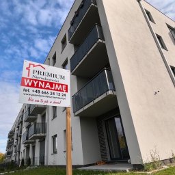 Premium R.E.A.Ltd-Artur Golabek - Nowoczesny budynek wielorodzinny z balkonami, przed którym umieszczono tablicę reklamową agencji nieruchomości z napisem 'Wynajmę' na tle błękitnego nieba.
