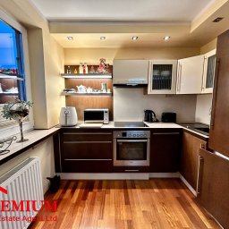 Premium R.E.A.Ltd-Artur Golabek - Nowoczesna kuchnia z drewnianą podłogą, brązowymi szafkami dolnymi i białymi górnymi, z widocznym oknem i dekoracjami na parapecie oraz logo agencji nieruchomości w lewym dolnym rogu.