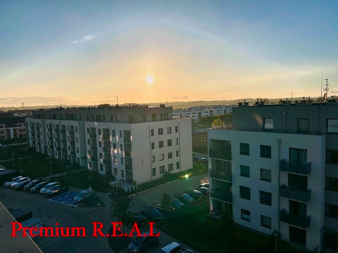 Widok z góry na osiedle mieszkaniowe o wschodzie słońca, z widocznymi budynkami, parkingiem i drzewami. Na zdjęciu widoczny napis 'Premium R.E.A.L.'