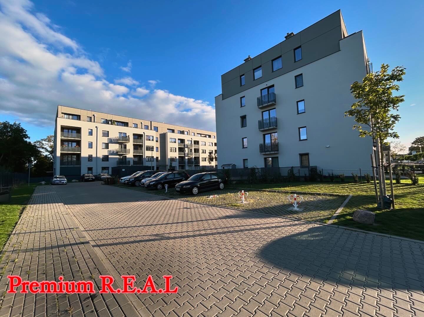 Nowoczesne budynki mieszkalne z balkonami, widok z parkingu wyłożonego kostką brukową, z logo firmy Premium R.E.A.L. w dolnej części kadru.