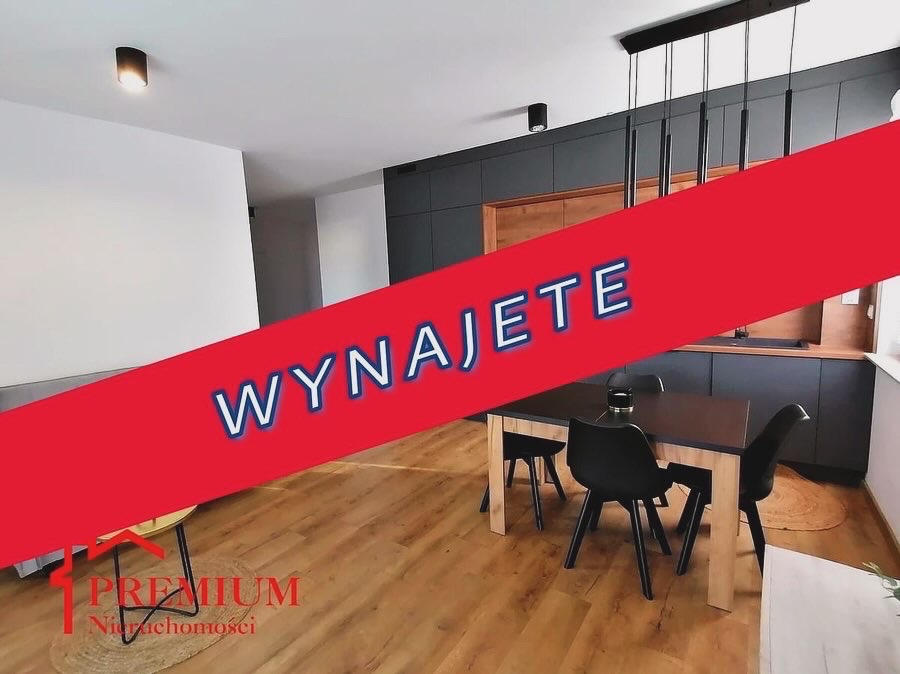 Jasne wnętrze mieszkania z drewnianą podłogą, czarnym stołem i krzesłami, oraz zabudową kuchenną w ciemnych odcieniach, przekreślone czerwonym banerem z napisem 'Wynajęte', w rogu logo agencji...